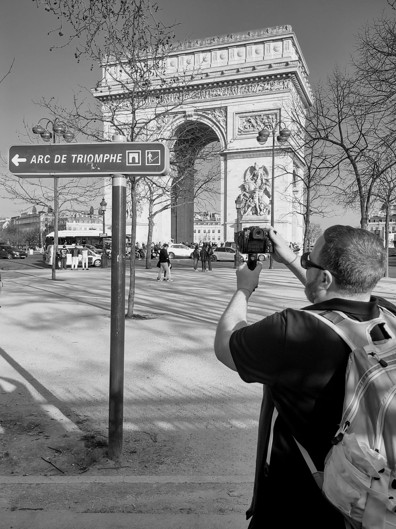 Tourist with backpack takes a photo of the Arc de Triomphe; foreground sign reads ARC DE TRIOMPHE with bare trees around.